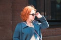 Attractive red-haired woman in a blue coat and jeans stands outside a brick building facade Royalty Free Stock Photo