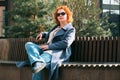 Attractive red-haired woman in a blue coat and sunglasses sits on a bench in a city block Royalty Free Stock Photo