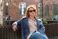 Attractive red-haired woman in a blue coat and sunglasses sits on a bench in a city block Royalty Free Stock Photo