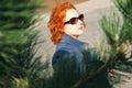 Attractive red-haired woman in a blue coat and sunglasses sits on a bench in a city block Royalty Free Stock Photo