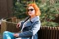 Attractive red-haired woman in a blue coat and sunglasses sits on a bench in a city block Royalty Free Stock Photo