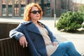 Attractive red-haired woman in a blue coat and sunglasses sits on a bench in a city block Royalty Free Stock Photo