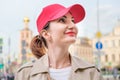 Young woman in a red baseball cap and beige raincoat stands against the background of city buildings and looks up