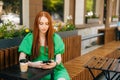 Attractive redhead young woman using browsing smartphone looking at screen, sitting at table with coffee cup in outdoor Royalty Free Stock Photo