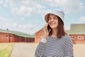 Attractive satisfied joyful dark haired woman wearing striped shirt and panama walking on streets talking mobile phone smiling