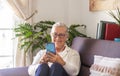 Attractive senior lady sitting on sofa at home using her smart phone for chatting with friends or family - woman white hair, Royalty Free Stock Photo