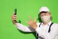 Attractive senior man with pink cap looking at camera wearing a fantasy medical mask due to covid-19 using smart phone app taking Royalty Free Stock Photo