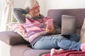 Attractive senior woman resting on sofa at home using laptop and mobile phone. Smiling elderly retiree enjoying tech and social Royalty Free Stock Photo