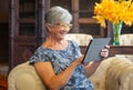 Attractive senior woman sitting on sofa using digital tablet. Smiling modern retiree enjoying relaxing moment