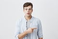 Attractive shocked young caucasian male with bugged eyes dressed casually pointing index finger sideways at grey blank