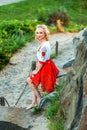 Attractive shoeless happy young blonde woman in stylish red white dress posing on path at hill. standing on road village and Royalty Free Stock Photo