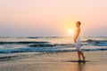 Attractive slender girl in white dress stands in waves on the sea in the light of setting sun. Young woman walks barefoot along Royalty Free Stock Photo