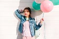 Attractive smiling girl in stylish denim jacket standing in shadow from colorful balloons in her hand. Charming young