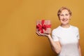 Attractive smiling senior woman in white t-shirt holding red gift box on bright yellow studio wall banner background Royalty Free Stock Photo