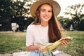 Attractive smiling teenage girl in straw hat holding corn in hands while happily looking in camera on picnic in city