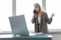 Attractive smiling young businesswoman wearing jacket talking on mobile phone while using laptop computer in office Royalty Free Stock Photo