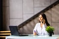 Attractive young businesswoman wearing jacket talking on mobile phone while sitting on a desk and using laptop computer in office Royalty Free Stock Photo