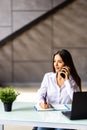 Attractive young businesswoman wearing jacket talking on mobile phone while sitting on a desk and using laptop computer in office Royalty Free Stock Photo