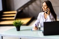 Attractive smiling young businesswoman wearing jacket talking on mobile phone while sitting on a desk and using laptop in office Royalty Free Stock Photo