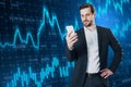 Attractive smiling young european businessman with cellphone and creative glowing forex chart on blurry background. Trade and Royalty Free Stock Photo