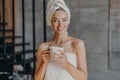 Attractive smiling young woman with makeup, toothy smile, poses bare shoulders, wrapped in towel, drinks hot beverage stands Royalty Free Stock Photo
