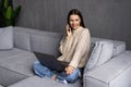 Attractive smiling young woman relaxing on a leather couch at home, working on laptop computer, talking on mobile phone Royalty Free Stock Photo