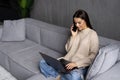 Attractive smiling young woman relaxing on a leather couch at home, working on laptop computer, talking on mobile phone Royalty Free Stock Photo