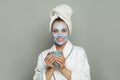 Attractive spa woman in cosmetic face mask drinking coffee in blue mug on gray background