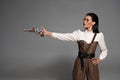 Steampunk woman standing with hand on hip and aiming with pistol on grey