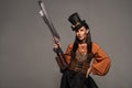 Steampunk woman in top hat holding gun and standing with hand on hip isolated on grey Royalty Free Stock Photo