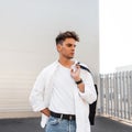 Attractive stylish young man in an elegant white shirt in blue jeans with a fashionable bag with a trendy hairstyle posing near a Royalty Free Stock Photo