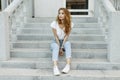 Attractive stylish young woman in a fashionable T-shirt in blue jeans in white trendy sneakers resting sitting on the steps Royalty Free Stock Photo