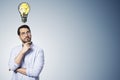 Attractive thoughtful young businessman with creative lamp sketch on gray wall background with mock up place. Idea, creativity and Royalty Free Stock Photo