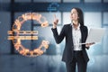 Attractive thoughtful young european businesswoman with laptop standing in blurry office interior with glowing euro sign. Online Royalty Free Stock Photo