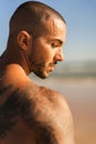 An attractive topless man with tattoos standing on a beach next to a sea