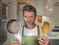 Attractive unhappy and overwhelmed home cook man in apron holding spoon and pan feeling upset and lazy at house kitchen in