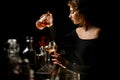 Attractive woman bartender in dark bar gently pours cocktail into wineglass.