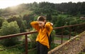 Attractive woman in casual clothes stands on a bridge in the mountains. Lady hiker in orange sweatshirt on the bridge on a