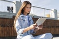 Attractive woman in denim clothes sits on park bench, holds smart tablet in her hands. Portrait of beautiful girl Royalty Free Stock Photo