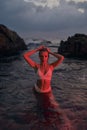 Attractive woman enjoys nocturnal swim in sea illuminated by crimson light. Nighttime ocean dip by stylish female