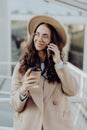 Attractive woman in eyeglasses talking on smart phone and holding paper cup of coffee in hand while standing outdoors. Trendy girl Royalty Free Stock Photo