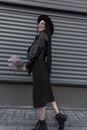 Attractive woman in fashion sunglasses in elegant hat in stylish black dress with bouquet of lilac poses near gray wall in street Royalty Free Stock Photo