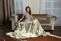 Attractive woman in fashionable silky shiny silver dress relaxing on brown sofa, fashion beauty portrait Royalty Free Stock Photo