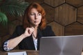 Attractive woman in a jacket nibbles thoughtfully at the temple of her glasses ends sitting in front of a laptop