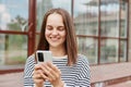 Attractive woman wearing striped shirt using mobile phone posing near building with big windows communicating with friends, typing