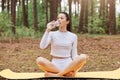 Attractive young adult woman wearing white sportswear sitting on kareman on ground, holding bottle and drinking water, feels