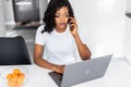 Attractive young african woman having phone conversation and smiling happily while doing paperwork at home, sitting at kitchen Royalty Free Stock Photo