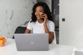 Attractive young african woman having phone conversation and smiling happily while doing paperwork at home, sitting at kitchen Royalty Free Stock Photo