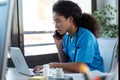 Attractive young afro female doctor talking with smart phone while working with her computer in the consultation Royalty Free Stock Photo