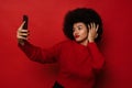 Attractive afro woman taking selfie on smartphone while standing isolated over red wall Royalty Free Stock Photo